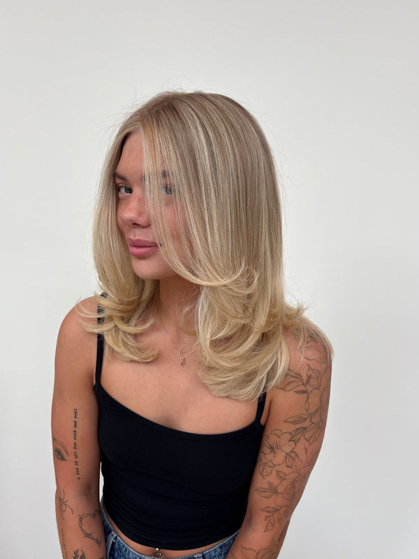 A woman with shoulder-length blonde hair wearing a black spaghetti-strap crop top and blue jeans, standing against a plain white wall. — after