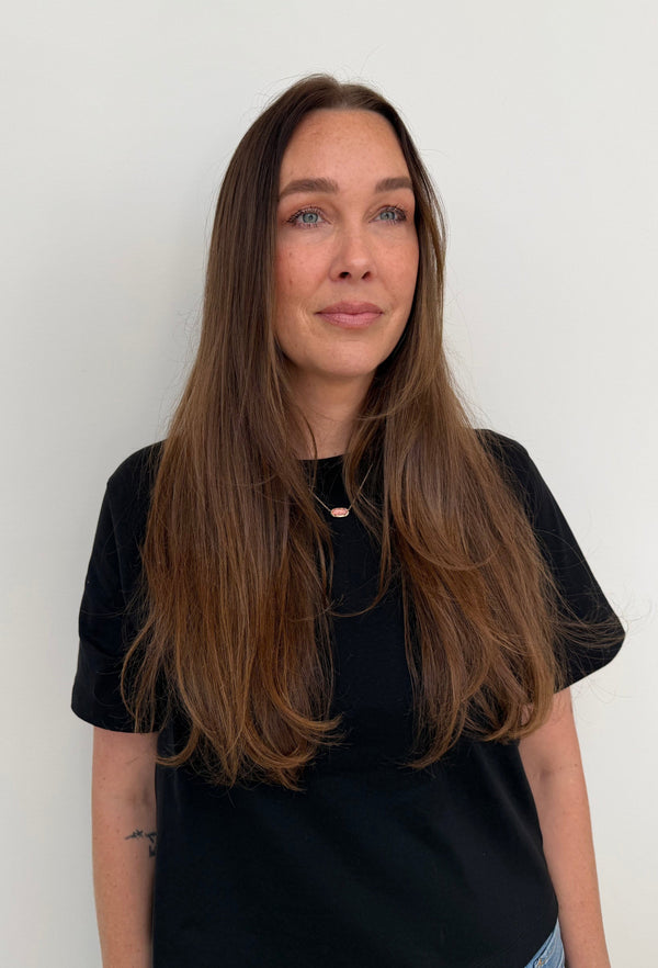 A woman with long brown hair wearing a black short-sleeve t-shirt and light blue jeans, standing against a plain white wall. — before