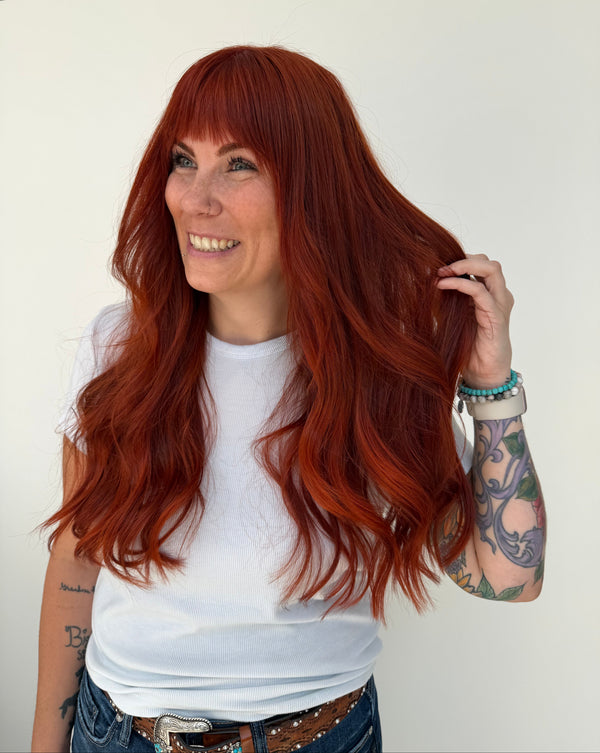 A woman with long, wavy, bright red hair wearing a white t-shirt and blue jeans, holding up a section of her hair to show the color and texture. She is standing against a plain white wall.